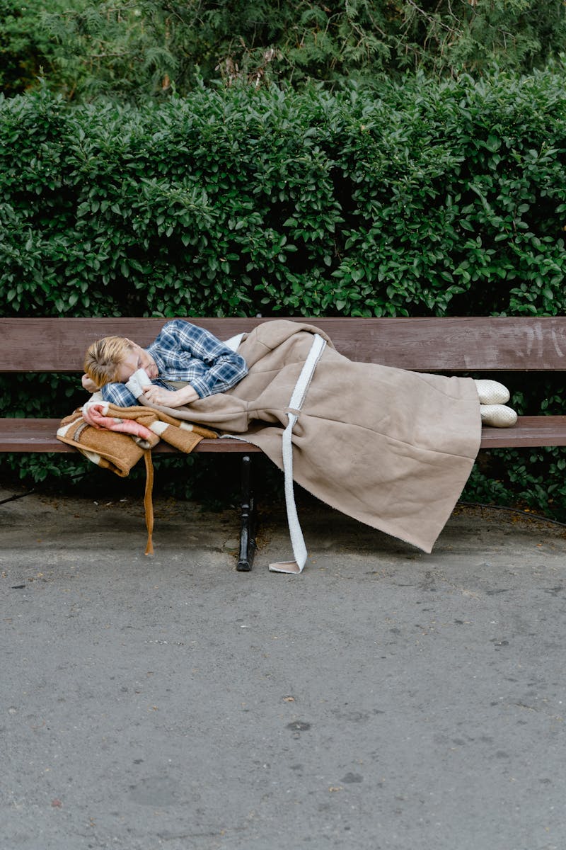 A woman sleeping wrapped in a blanket on a park bench, illustrating homelessness and solitude.