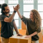 Happy woman in casual wear standing near heap of cardboard boxes and giving high five to ethnic boyfriend with ponytail showing agreement while looking at each other